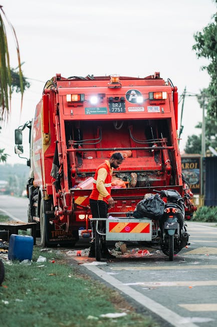 A municipal waste collection worker in high-visibility orange and yellow clothing is seen operating a large, red rubbish collection vehicle on a paved urban street. The back of the vehicle is open, revealing internal mechanical components and a tilted loading area. The worker is actively engaged in emptying a black rubbish bag into the vehicle's hopper, with a small black trash bin positioned nearby on a metal stand. The scene includes a partial view of a motorbike parked close to the worker, and nearby the sidewalk is lined with green grass, trees, and utility poles with electrical wires. In the background, there are commercial signs and buildings, indicating a busy street environment. The lighting appears natural, possibly overcast, providing even illumination across the area. This scene illustrates an on-site waste collection process, exemplifying the services provided by companies like Rubbish Removal Bow that offer alternative waste handling options for urban environments, bypassing traditional council bin collections.