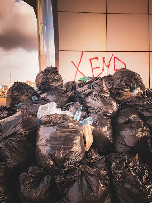A large pile of black plastic rubbish bags is stacked against a modern tiled wall, with some partly torn or stretched, revealing white and brown waste inside. Several clear plastic bottles are visible protruding from the bags, indicating discarded bottles among the waste. Behind the bags, there is graffiti with the word 'XEND' spray-painted in red on the wall, which is composed of beige tiles with visible grout lines. The scene appears outdoors, possibly on a pavement or alleyway, with natural lighting suggesting late afternoon or early evening, and a cloudy sky visible in the background. The presence of the bags and bottles implies an area where waste has been temporarily accumulated, potentially awaiting collection or disposal, aligning with the context of independent rubbish removal or alternative waste handling services as performed by companies like Rubbish Removal Bow.