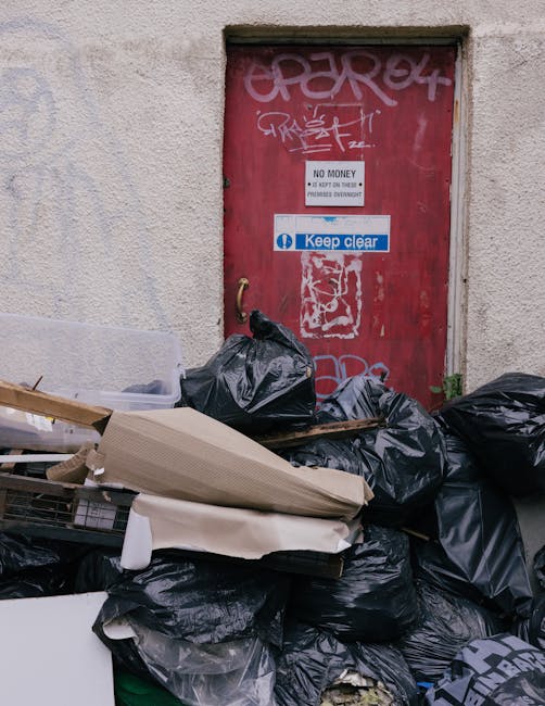 A municipal waste collection worker in high-visibility orange and yellow clothing is seen operating a large, red rubbish collection vehicle on a paved urban street. The back of the vehicle is open, revealing internal mechanical components and a tilted loading area. The worker is actively engaged in emptying a black rubbish bag into the vehicle's hopper, with a small black trash bin positioned nearby on a metal stand. The scene includes a partial view of a motorbike parked close to the worker, and nearby the sidewalk is lined with green grass, trees, and utility poles with electrical wires. In the background, there are commercial signs and buildings, indicating a busy street environment. The lighting appears natural, possibly overcast, providing even illumination across the area. This scene illustrates an on-site waste collection process, exemplifying the services provided by companies like Rubbish Removal Bow that offer alternative waste handling options for urban environments, bypassing traditional council bin collections.