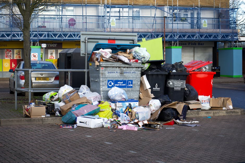 A large collection of discarded waste is piled on a paved area in front of a commercial building, including open and closed black and red rubbish bins, filled with mixed waste such as cardboard boxes, plastic bags, paper, and packaging materials. The waste extends onto the surrounding pavement, with some items spilling onto the brick surface, and additional cardboard boxes and plastic sacks placed around the bins. In the background, there is a parked silver car with a visible yellow license plate, and the building behind features a storefront with signage and closed roller shutters, indicating a retail or food establishment. The scene is outdoors, with a leafless tree on the left and a protective metal railing bordering the waste area. The overall environment suggests a typical urban or commercial setting where private rubbish disposal or alternative waste collection methods might be employed, aligning with the services offered by Rubbish Removal Bow.