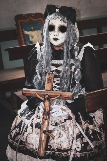 A person dressed in a gothic-style costume sits indoors on a dark wooden bench, holding a wooden cross with a small figure affixed to it. The costume features a long, black dress with white lace and patterned fabric, and includes black puffed sleeves, and an intricate design that resembles medieval or Victorian fashion. Their hair is long, silver-gray, styled in loose waves, and they wear a large black bow on top of their head. The person's face is painted pale with dark eye circles and dark lipstick, and they have black circles around their eyes, contributing to a ghostly or skull-like appearance. In the background, there is a religious icon or painting of Jesus with open arms, mounted on an aged wall, which contrasts with the eerie costume. The setting appears to be a dimly lit, indoor environment, possibly a church or a room decorated with religious artifacts, with the overall atmosphere conveying a sense of solemnity and alternative cultural expression related to gothic or ritualistic themes.