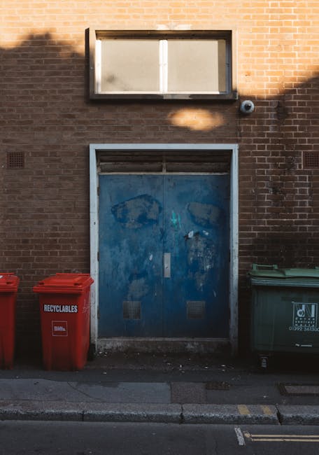 A large collection of discarded waste is piled on a paved area in front of a commercial building, including open and closed black and red rubbish bins, filled with mixed waste such as cardboard boxes, plastic bags, paper, and packaging materials. The waste extends onto the surrounding pavement, with some items spilling onto the brick surface, and additional cardboard boxes and plastic sacks placed around the bins. In the background, there is a parked silver car with a visible yellow license plate, and the building behind features a storefront with signage and closed roller shutters, indicating a retail or food establishment. The scene is outdoors, with a leafless tree on the left and a protective metal railing bordering the waste area. The overall environment suggests a typical urban or commercial setting where private rubbish disposal or alternative waste collection methods might be employed, aligning with the services offered by Rubbish Removal Bow.