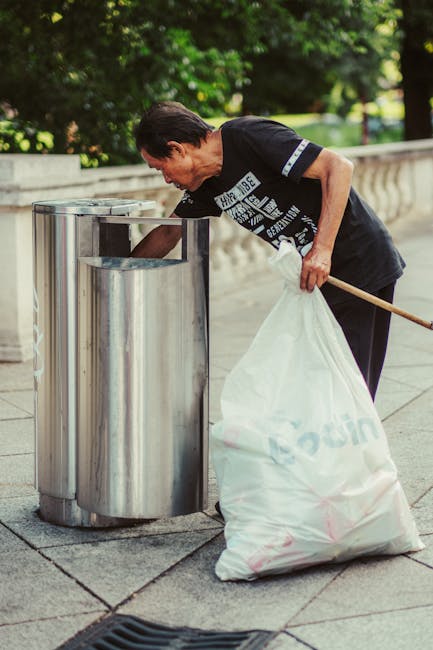 A middle-aged woman with dark hair tied back is seen on a paved sidewalk near a stone balustrade, disposing of rubbish into a stainless steel public bin. She is wearing a black short-sleeved T-shirt with printed text on the front, black trousers, and appears focused on emptying a large, semi-transparent white plastic bag filled with waste. The bag, marked with a printed logo, is held in her left hand, while her right hand is reaching into the bin to deposit the rubbish. The surrounding environment includes lush green trees and neatly trimmed grass in the background, suggesting a park or urban outdoor space. The scene is well-lit with natural daylight, casting soft shadows on the ground. This image exemplifies a private or community-led rubbish disposal action aligned with waste management services such as those provided by Rubbish Removal Bow, highlighting the importance of proper waste handling in public areas.