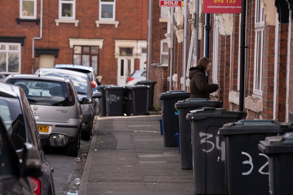 A woman standing on a sidewalk beside a row of large, dark grey wheelie bins with hinged lids, positioned in front of a brick residential building. The bins are arranged in a line along the pavement, with some showing chalk markings or graffiti. The woman, wearing a brown jacket with a hood, is engaged in an activity near the first bin, possibly disposing of waste or organizing the bins. To her right, the building features white window frames and a red advertising sign, partially visible, indicating a property for sale. The street behind her is lined with parked cars, including a silver hatchback closest to the camera, and extends into the background where additional cars and more brick terraced houses can be seen. The environment appears to be in a residential area with an urban street scene, typical of the type of location serviced by Rubbish Removal Bow, highlighting a setting where on-site waste management or private waste collection services are relevant.