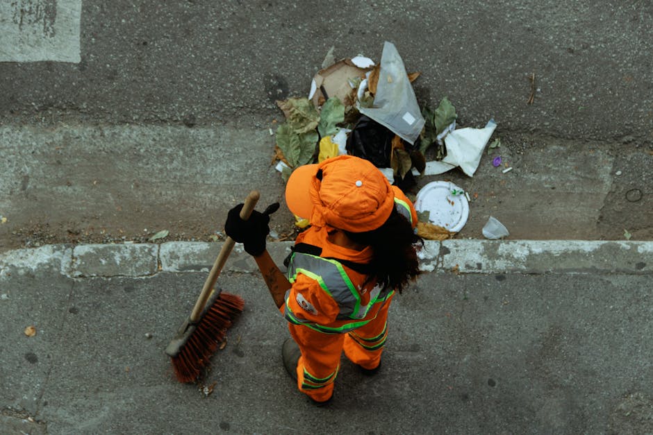 A waste collection worker dressed in high-visibility orange safety gear with reflective stripes, wearing a matching orange cap, is seen from above while standing on a concrete pavement. The worker is holding a long-handled broom and appears to be in the process of clearing litter from a pile of waste materials, including crumpled paper, plastic packaging, and food waste, which is situated against a concrete wall or curb. The waste is scattered across the edge of the pavement, with some debris spilling onto the street surface. The scene is outdoors, with natural daylight illuminating the area. The worker’s posture and the tools suggest an on-site clearance operation typical of private rubbish removal services like those offered by Rubbish Removal Bow, providing an alternative to municipal waste collection, tailored for efficient debris management in urban settings.