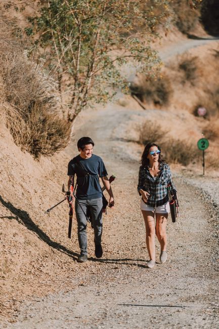 A man and a woman are walking along a gravel path in a dry, desert-like outdoor environment. The man is wearing a dark T-shirt, grey pants, and gloves, carrying a pair of safety tools or equipment in his hands. The woman is dressed in a checked shirt, beige shorts, and sunglasses, holding a clipboard or folder in one hand. The background features sparse bushes, small trees, and rocky hills with a warm, sunlit atmosphere. To the right of the pair, a green numbered trail marker is visible, indicating a designated walking route. The scene evokes a sense of outdoor exploration or inspection, potentially related to environmental or site management activities, which could be connected to private waste handling or site clearance services offered by Rubbish Removal Bow. The natural terrain and the individuals' purposeful movement suggest on-site activities that might include environmental assessments or land management, aligning with the theme of alternative waste or debris removal services outside conventional rubbish collection methods.