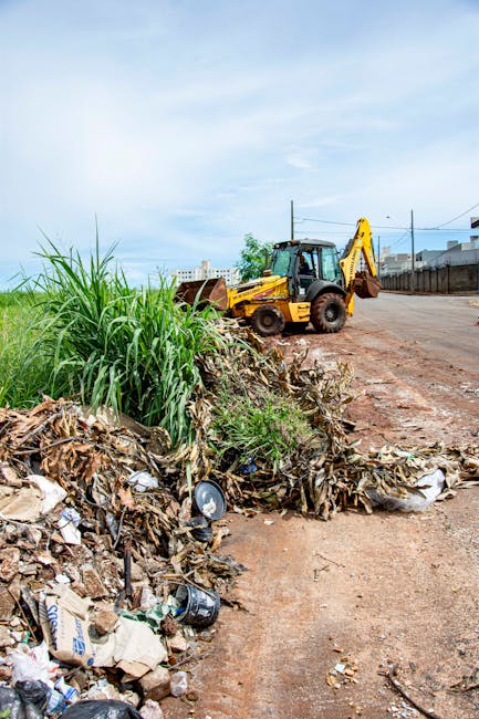 A waste collection worker dressed in high-visibility orange safety gear with reflective stripes, wearing a matching orange cap, is seen from above while standing on a concrete pavement. The worker is holding a long-handled broom and appears to be in the process of clearing litter from a pile of waste materials, including crumpled paper, plastic packaging, and food waste, which is situated against a concrete wall or curb. The waste is scattered across the edge of the pavement, with some debris spilling onto the street surface. The scene is outdoors, with natural daylight illuminating the area. The worker’s posture and the tools suggest an on-site clearance operation typical of private rubbish removal services like those offered by Rubbish Removal Bow, providing an alternative to municipal waste collection, tailored for efficient debris management in urban settings.