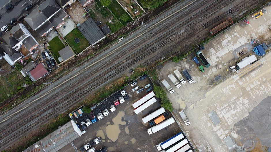 An aerial view showing a collection of various white and grey delivery and storage trucks, trailers, and containers parked on a paved lot adjacent to train tracks. The trucks are positioned along the right side of the image, with some parked parallel and others at angles. Puddles and patches of water are visible on the concrete surface, indicating recent rain or poor drainage. To the left, there are small residential houses with rooftops of different materials, including slate and tile, some with small gardens or yards. The train tracks run horizontally across the upper part of the image, separated from the parking area by a strip of greenery and weeds. The scene appears to depict a commercial or industrial area where waste or bulk goods might be collected and stored, aligning with private waste disposal services such as those offered by Rubbish Removal Bow nearby. The overall atmosphere is utilitarian, with a focus on the transportation and storage of large objects and materials within an urban environment.