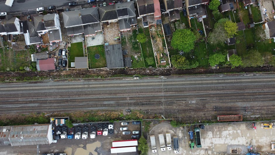 An aerial view showing a row of residential houses with small gardens and yards located behind a set of railway tracks. The backyards vary in size and features, including grass lawns, paved areas, sheds, and small patios. Some gardens have visible trees and shrubs, contributing to a green and lively environment. Vehicles are parked along the street and within adjacent parking areas, with a variety of cars and vans visible. In the foreground, there is a small collection of discarded rubbish, including plastic containers, cardboard boxes, and other debris, situated on a paved surface next to a building. The scene appears to depict a typical urban setting where private waste collection or rubbish removal services, like those provided by Rubbish Removal Bow, might be involved in clearing waste from residential or commercial properties, often separate from public authority disposal systems. The overall lighting suggests daytime with overcast skies, and the environment is clean and organized aside from the waste area, supporting the context of professional rubbish removal and on-site clearance activities.