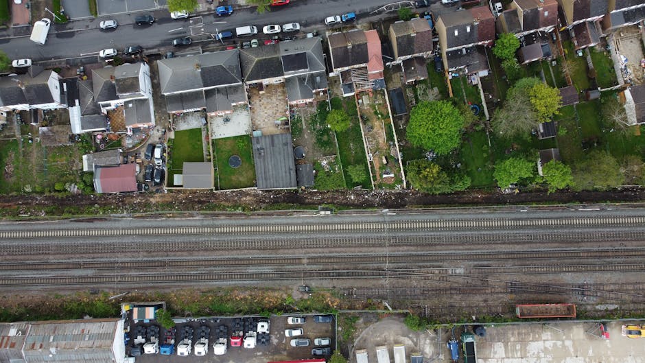 An aerial view showing a collection of various white and grey delivery and storage trucks, trailers, and containers parked on a paved lot adjacent to train tracks. The trucks are positioned along the right side of the image, with some parked parallel and others at angles. Puddles and patches of water are visible on the concrete surface, indicating recent rain or poor drainage. To the left, there are small residential houses with rooftops of different materials, including slate and tile, some with small gardens or yards. The train tracks run horizontally across the upper part of the image, separated from the parking area by a strip of greenery and weeds. The scene appears to depict a commercial or industrial area where waste or bulk goods might be collected and stored, aligning with private waste disposal services such as those offered by Rubbish Removal Bow nearby. The overall atmosphere is utilitarian, with a focus on the transportation and storage of large objects and materials within an urban environment.